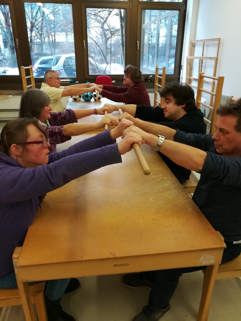 During a Conductive Session: Six adults are sitting at a table opposite each other building pairs. Each opposite pair hols onto an excercise stick either side. Everyone sits on Petö Chairs.