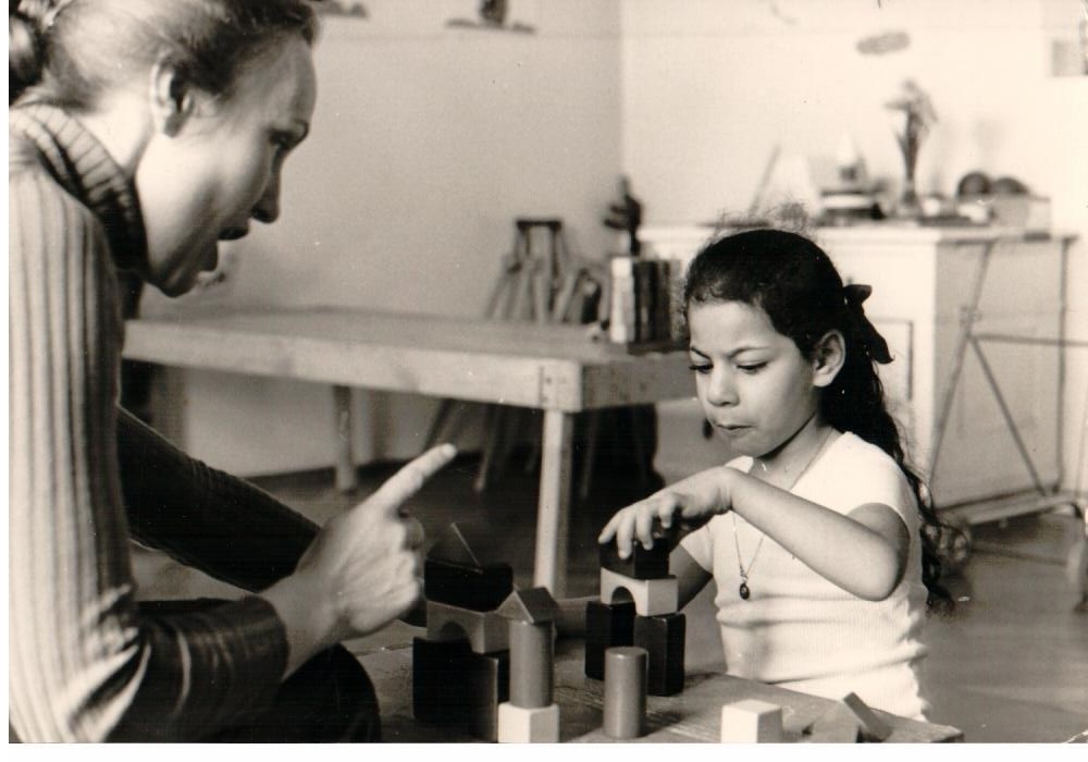 black and white picture. Teacher sitting with child at table talking to the girl, pointing a finger. The girl piles up bricks to a house.