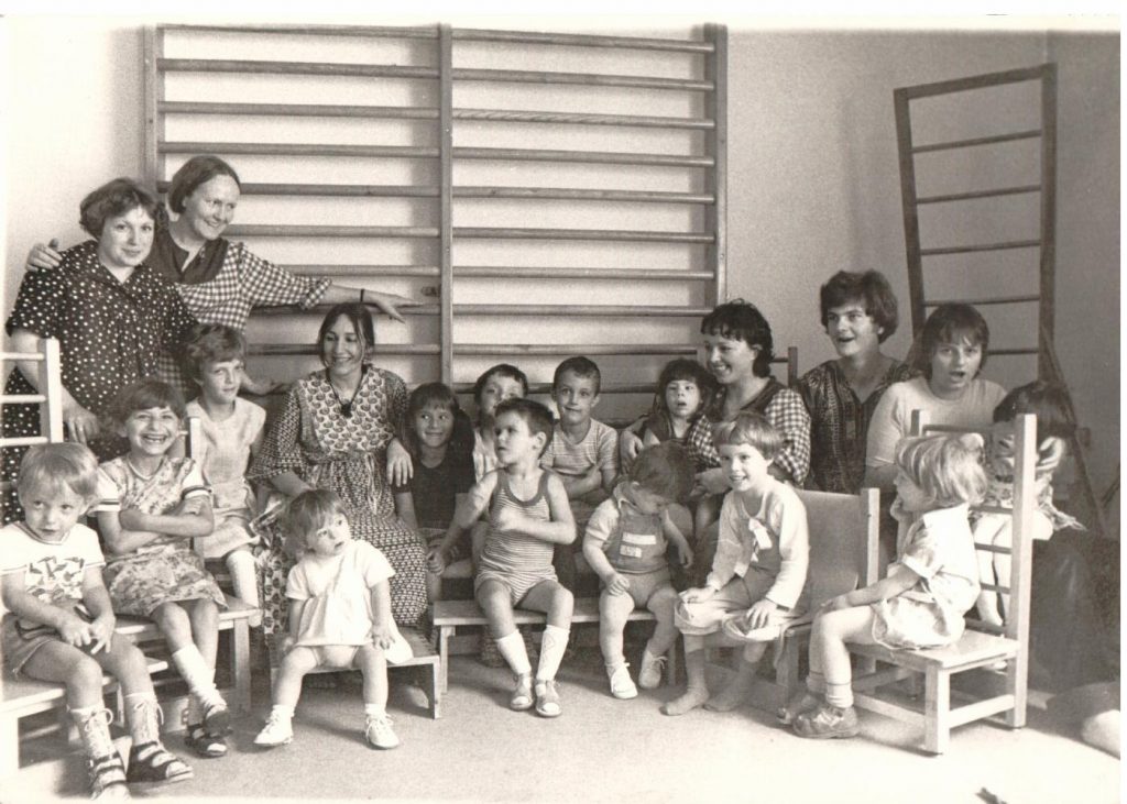 black and white: Children sitting on chairs with teacher. Group picture. In the back ground wall ladders.