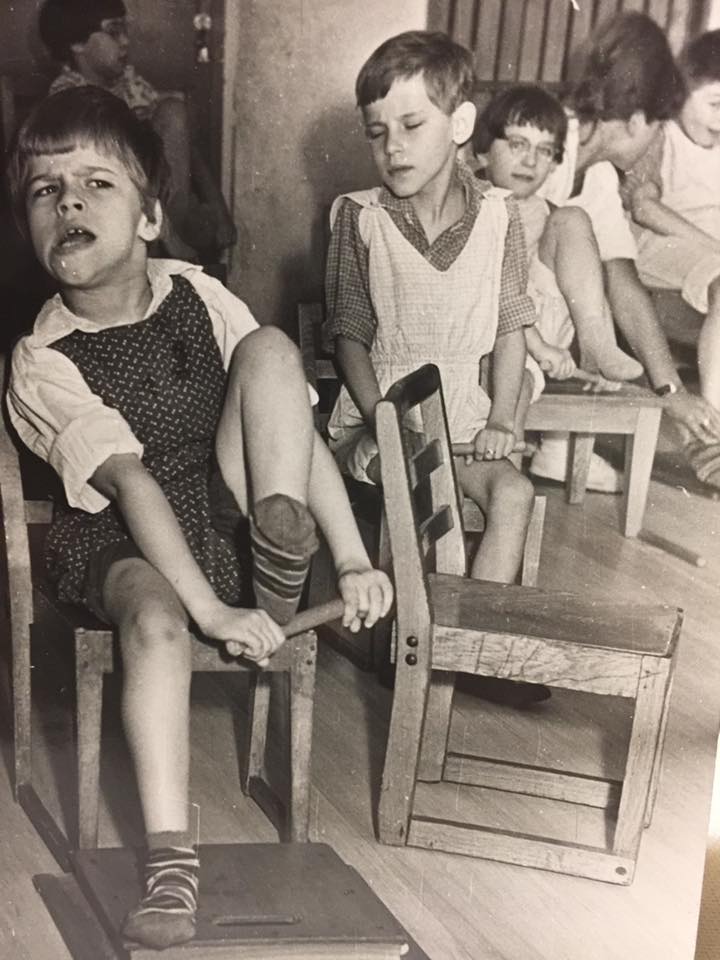 Black, white: small children sitting on wooden chairs during CE excercise. They try to pull their leg up and lift it over a stick which they are holding with two hands.